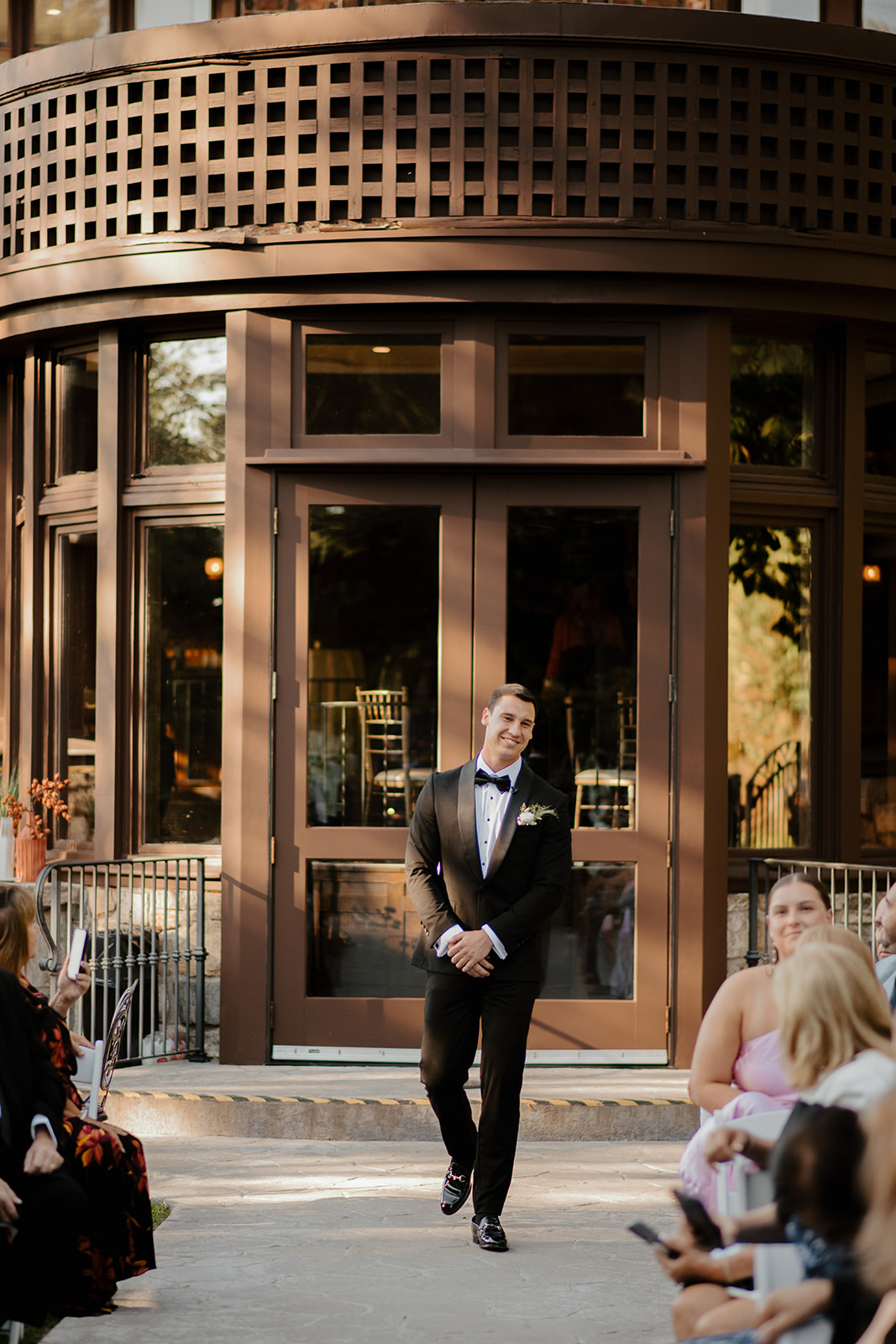 Groom in a tuxedo walking down the outdoor ceremony aisle with a smile as guests watch.